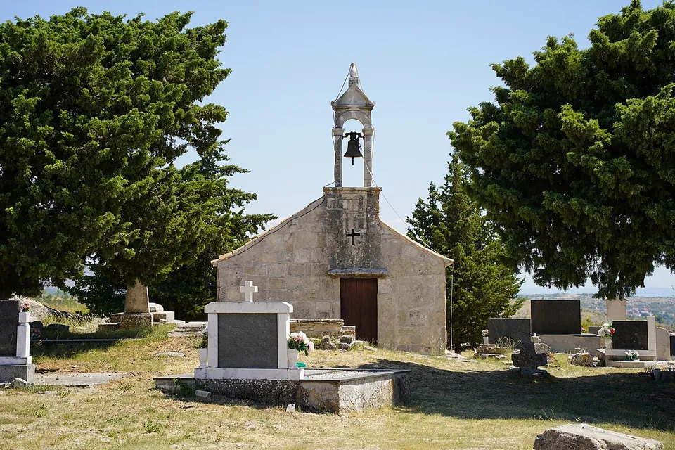 A small stone chapel with a single bell in an open bell-cote, among cypress trees and gravestones in a small cemetery.