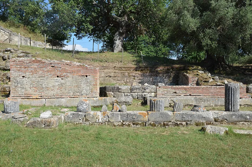 Low walls of brick and stone forming a square building on a lawn, with column drums lying and standing in front and oak trees behind.