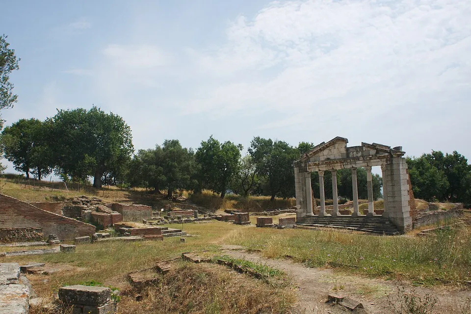 A grassy archaeological site with scattered stone blocks, and at the back a re-erected building front of six columns carrying an entablature and pediment, with olive trees behind.