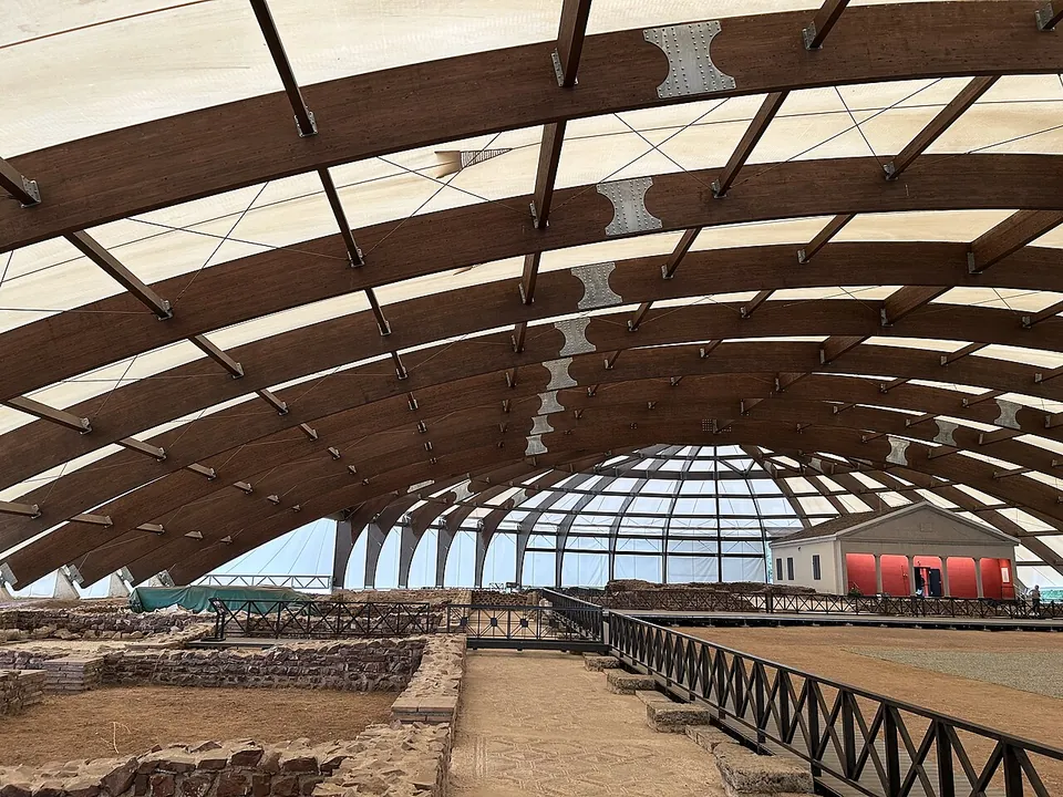 The inside of a large modern canopy of curved timber ribs and pale fabric, spanning the excavated walls of the villa, with a raised walkway along one side and the red-panelled pavilion visible beyond the open end.