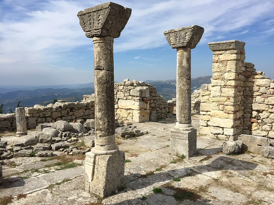 The curved apse wall of a church and rows of column bases and a few standing columns in a green meadow, under a clear sky.