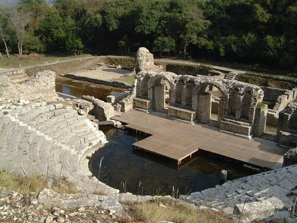 A theatre's curved stone seating dropping to a flooded orchestra, with a wooden walkway laid over the water in front of the ruined stage building and its row of arches, among trees.