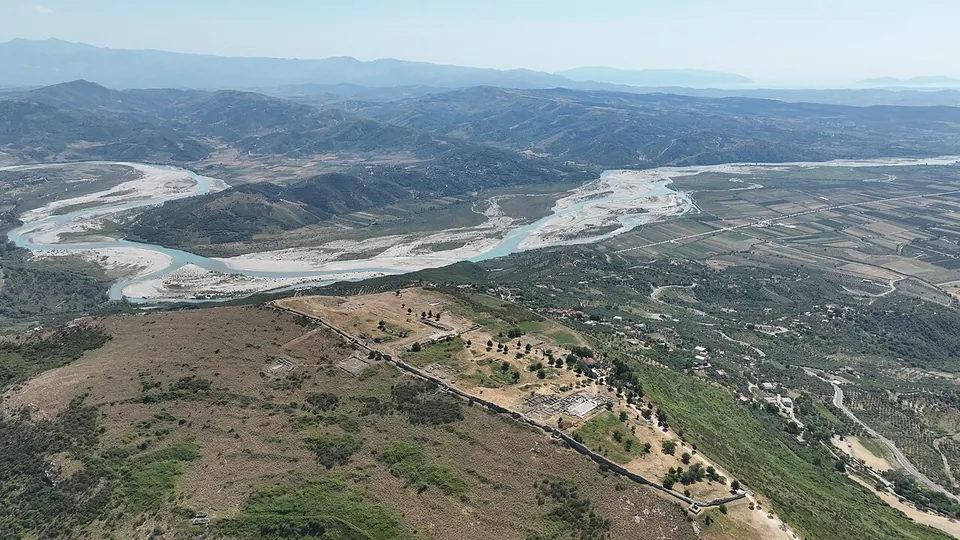 An aerial view of a long hilltop with the line of an ancient city wall and scattered ruins, above a broad braided river valley and ranges of hills.