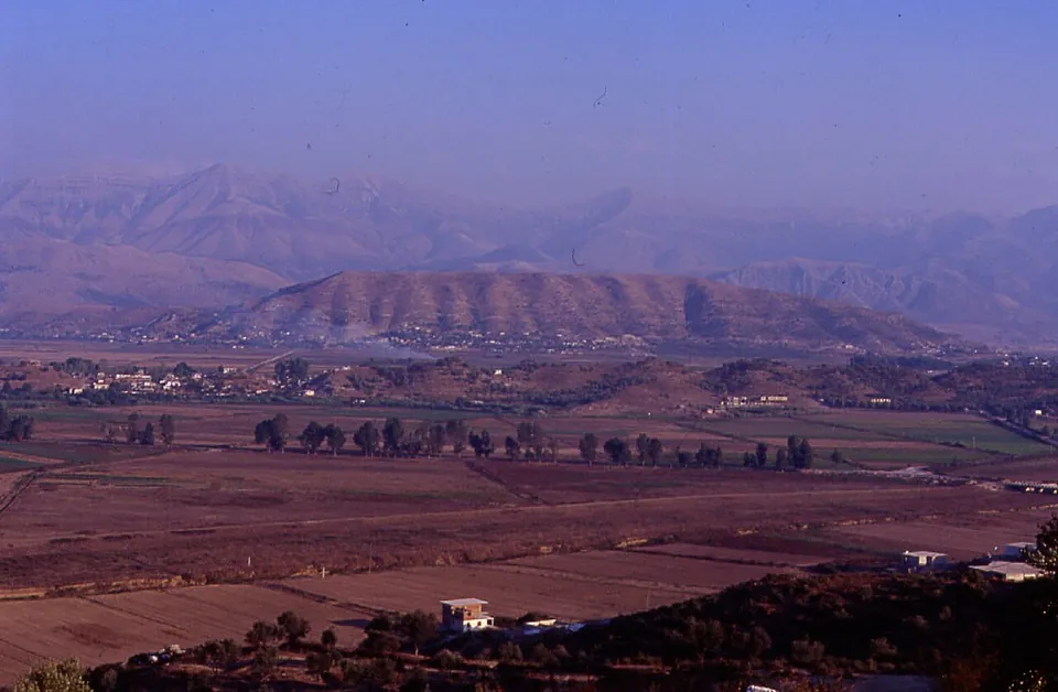 A long bare hill rising above a plain of fields and trees, with higher mountains behind in a hazy light.