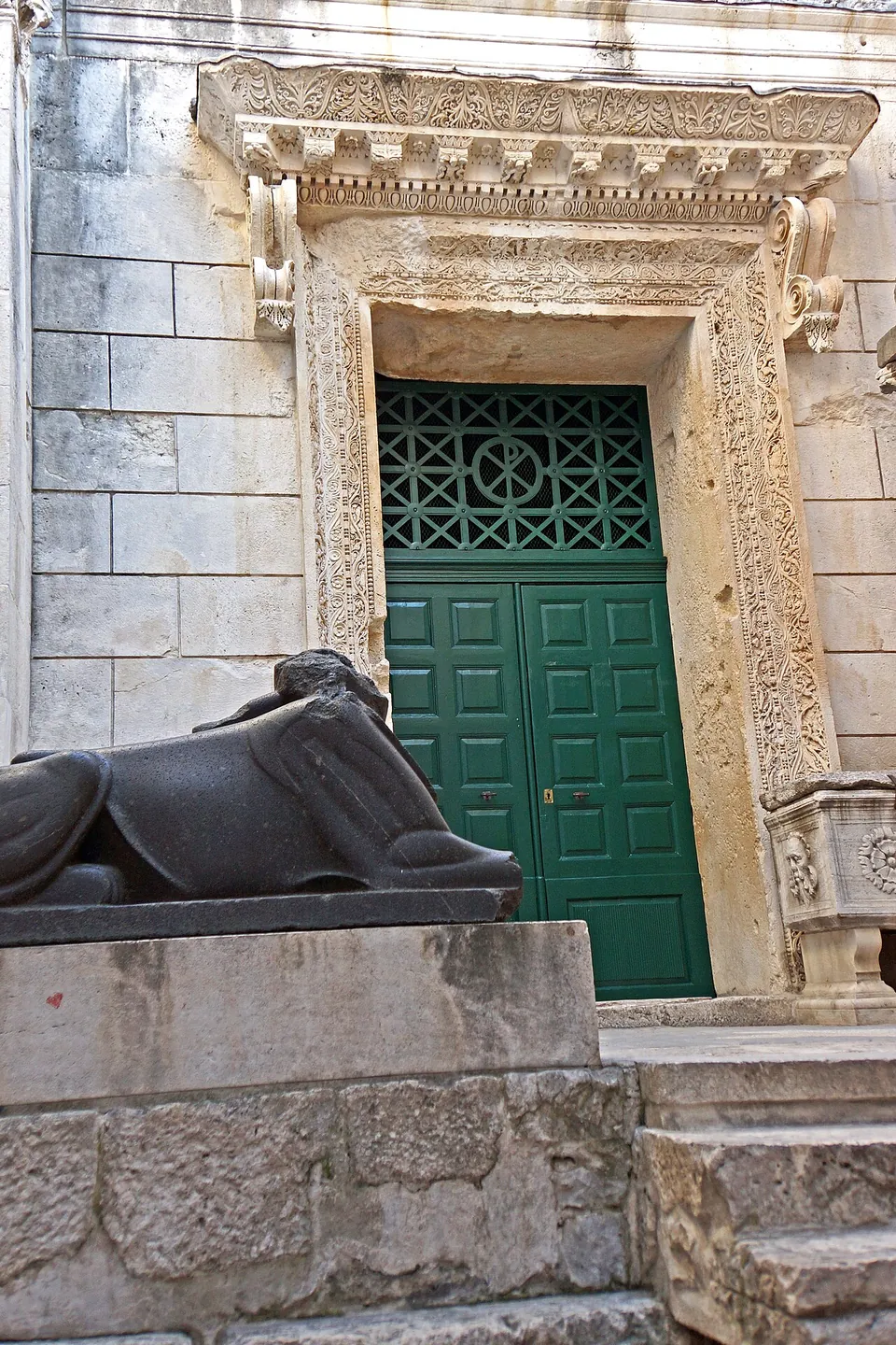 A doorway with a deeply carved stone frame and green doors at the top of a few steps, with a headless black granite sphinx lying on a stone base in front of it.