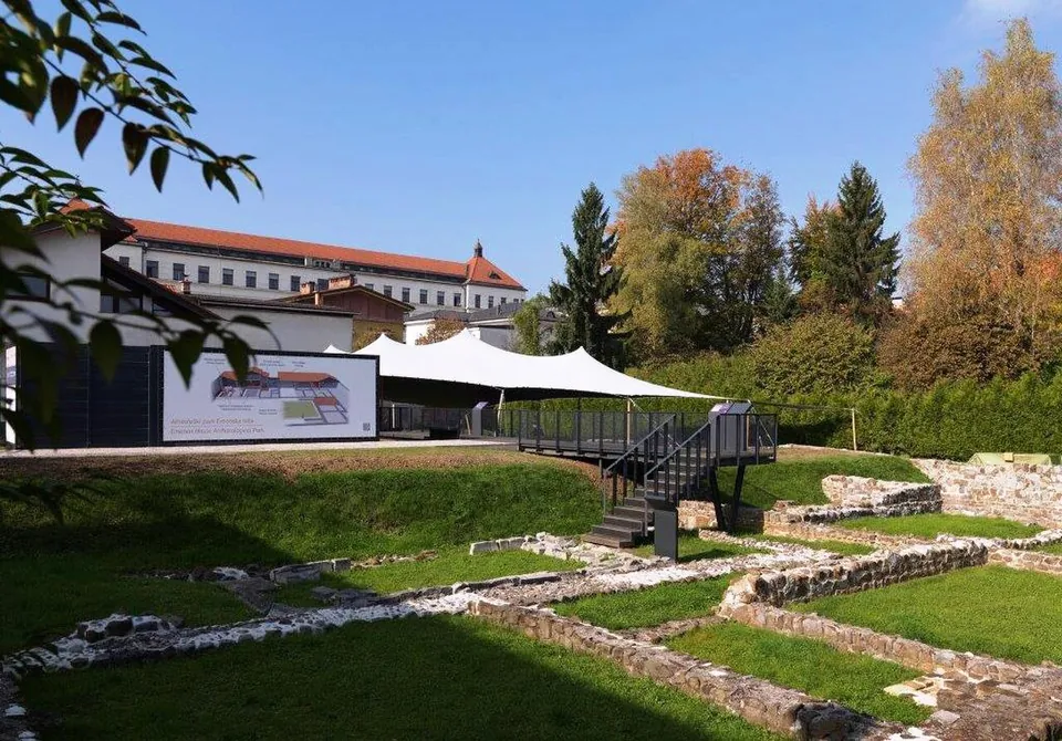 A sunken lawn crossed by low excavated stone walls, with a black metal viewing platform and steps, a white fabric canopy behind it, an illustrated information panel on the left and a large red-roofed building beyond the trees.