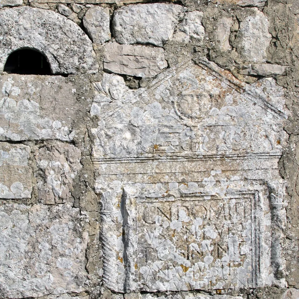 A weathered Roman relief set into a rough stone wall, a pediment with a carved head above a framed panel with a worn inscription.