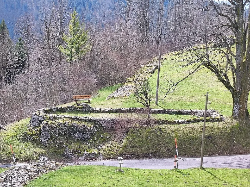 Low stone foundations of a small rectangular building on a grassy terrace above a road, with a wooden bench beside them and bare trees on the slope behind.
