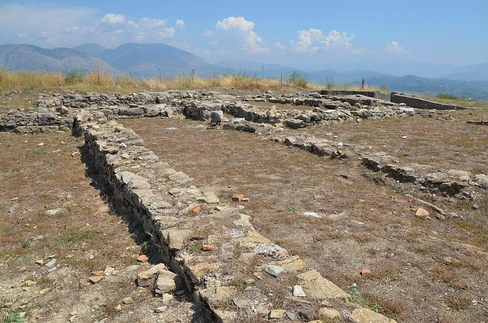 Standing stone pillars and column drums among low walls of large blocks on a hilltop, with a wide valley and mountains beyond.