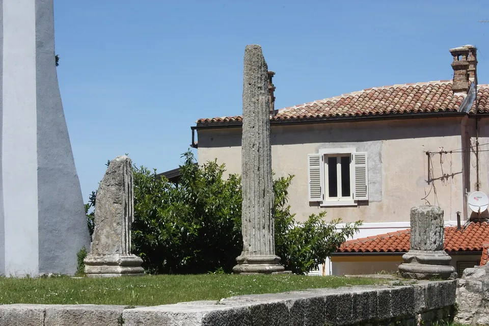 Two tall weathered stone column shafts and a short fragment standing on a grassy terrace in front of a house with shutters and a red-tiled roof.