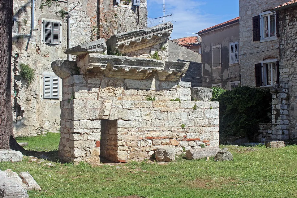 The ruined corner of a Roman temple built of large squared stone blocks, carrying fragments of a carved cornice, standing on a lawn among old stone houses.