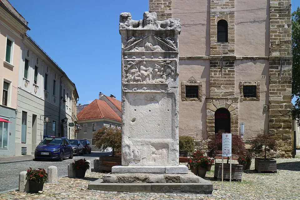 A tall weathered marble stele standing on a cobbled square, carved with worn relief panels and crowned by lions, with a pale church tower behind it and townhouses along the street to the left.