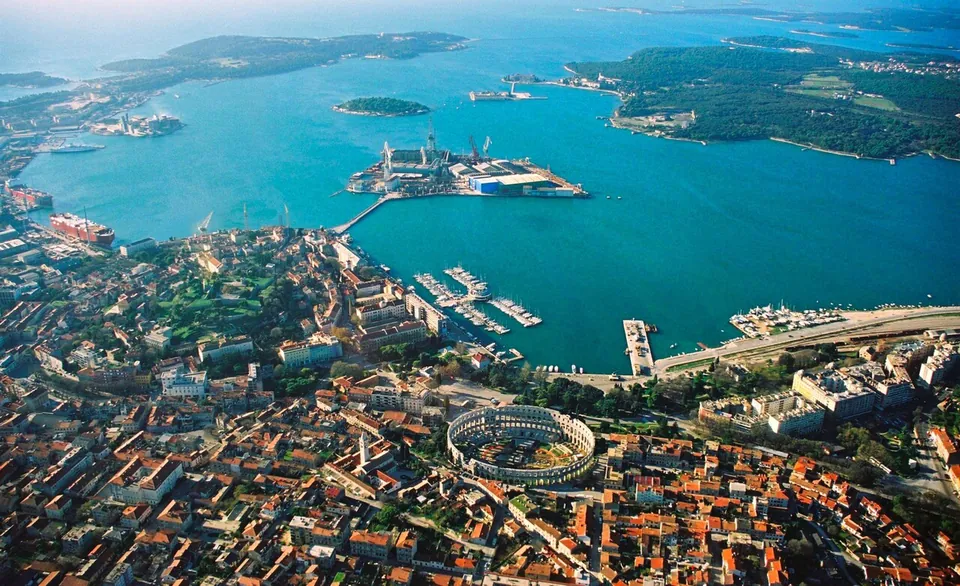 An aerial view of a coastal town of red roofs around a wide blue bay, with a shipyard on an island in the harbour, wooded islands beyond, and an oval Roman amphitheatre standing out among the buildings in the foreground.
