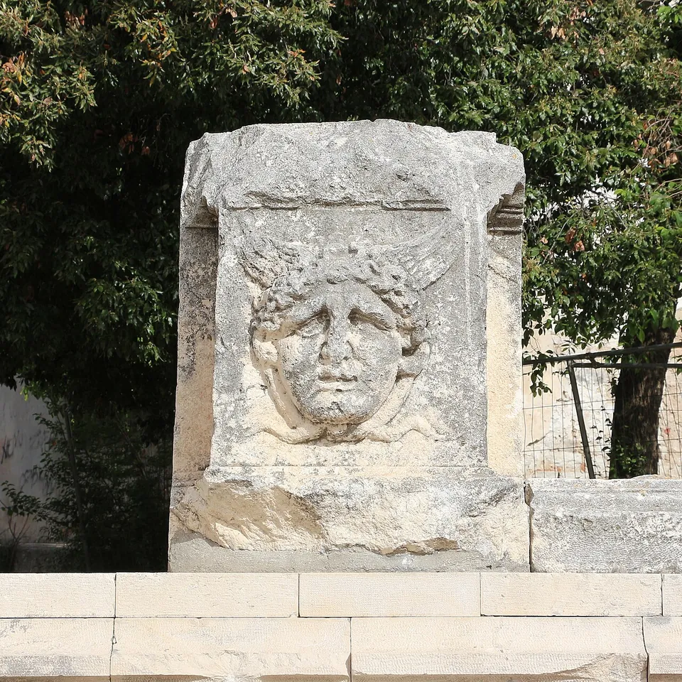 A worn stone block carved with a round face framed by wild hair, set on a stone base against dark foliage.