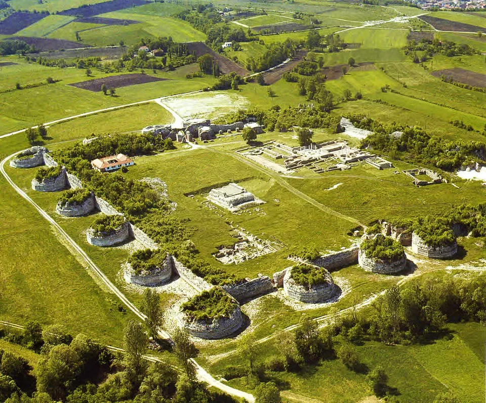 An aerial view of the whole site, an irregular ring of massive round towers and connecting wall enclosing mown grass, with the excavated foundations of the palace and its buildings laid out inside and farmland beyond.