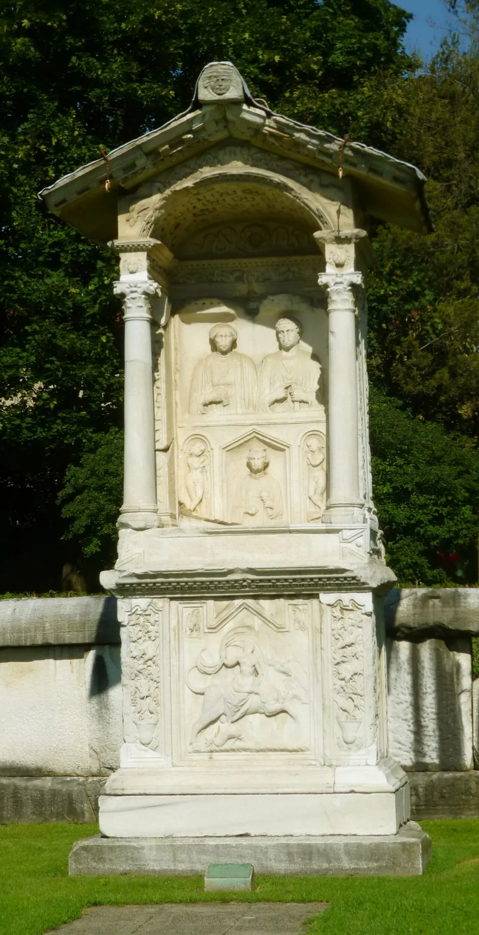A white marble tomb shaped like a small shrine, with two portrait busts in a niche between columns under a pitched roof, and a large relief of a figure and an animal on the base below.