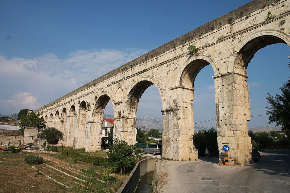 A tall arcade of pale limestone arches on massive piers, seen from below at an angle, with a narrow road passing under the nearest arch and a road sign at its foot.