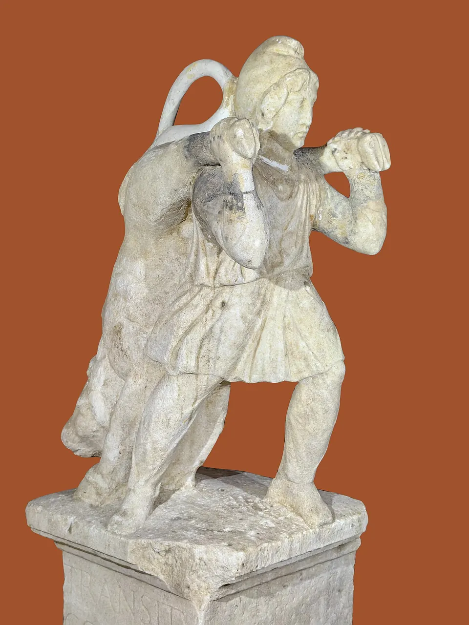 A marble statue of a man in a short tunic and cloak, striding forward, bent under the weight of a bull slung across his shoulders, on a squared base; the background is plain reddish brown.
