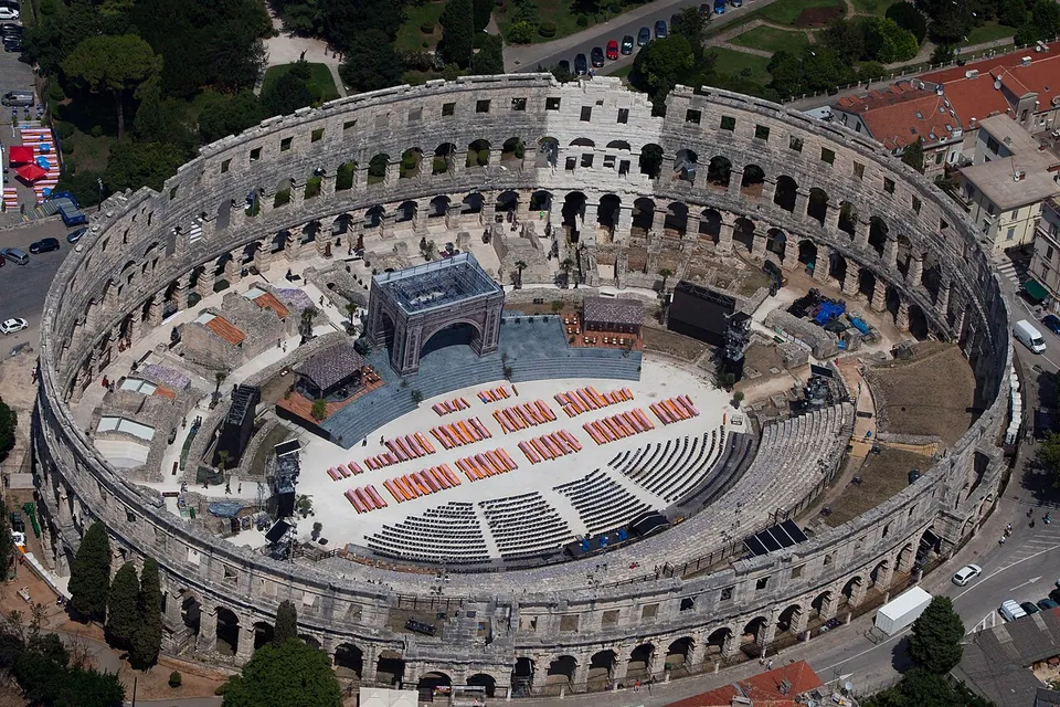 An aerial view of an oval Roman amphitheatre with its outer wall of arches standing complete, the arena floor inside filled with a modern stage and rows of red seats, surrounded by town streets and trees.
