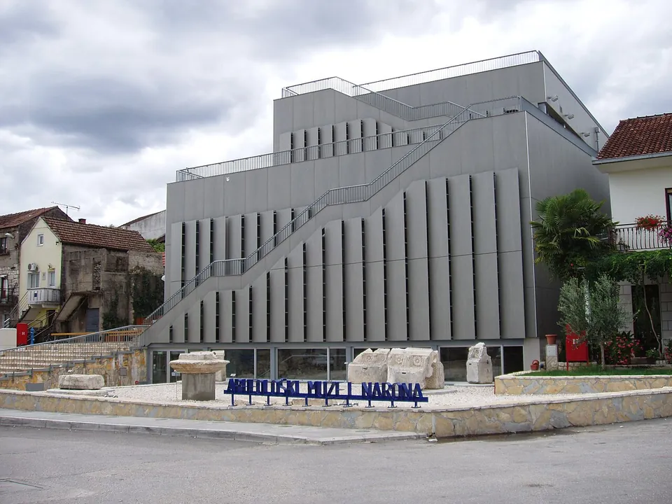 A modern grey museum building with a diagonal external staircase, stone fragments on its forecourt and large blue letters reading "Arheološki muzej Narona" at the entrance.